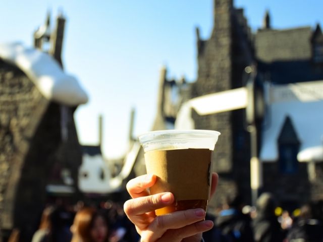 Hand holding a full cup of Butterbeer with towering snowcapped Hogwarts castle in the background. Keep reading to to find out more Mistakes to Avoid at Universal Orlando Resort!