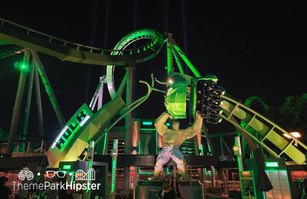 Hulk Roller Coaster Entrance glowing green at Universal Orlando Resort Islands of Adventure with the Hulk holding the coaster track over his head. Keep reading to find out more about Hulk vs VelociCoaster at Universal Orlando.