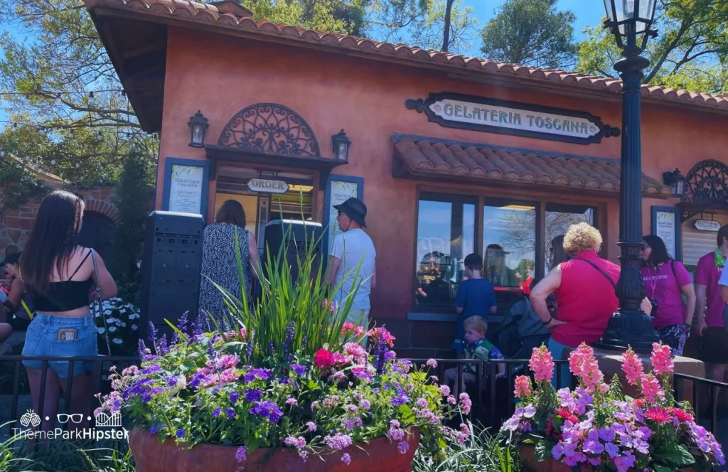 Italy Pavilion Gelateria Toscana  in Epcot during the Flower and Garden Festival 2023 decorated with brightly colored flowers in planters while guests stand outside. Keep reading to find out the must-try best snacks at EPCOT. 