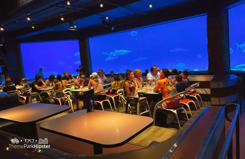 SeaWorld Orlando Resort Sharks Underwater Grill dining area with tables for guests in front of an entire wall of panoramic views of the aquarium. Keep reading to learn more about Sharks Underwater Grill at SeaWorld Orlando.