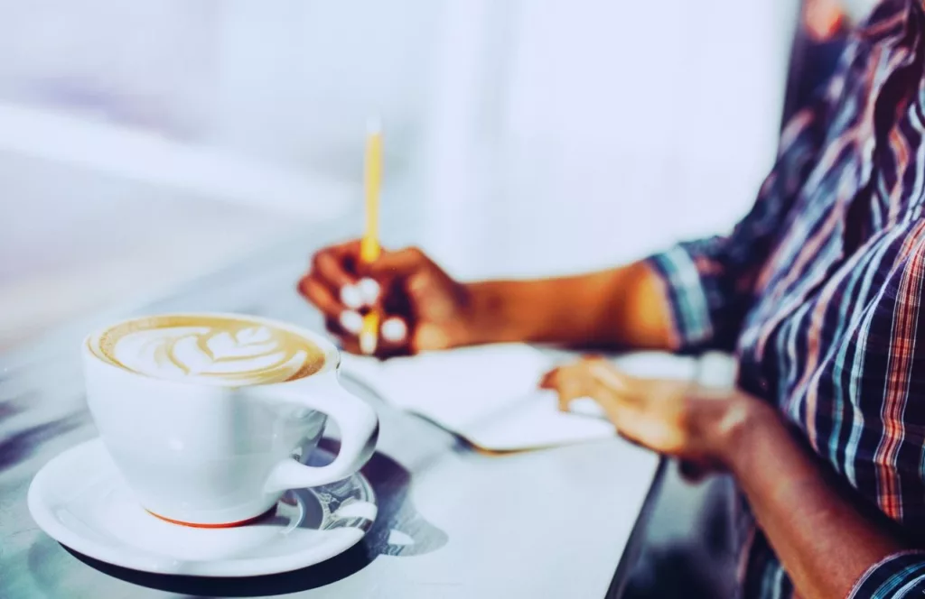 Black woman journaling on vacation next to coffee. Keep reading to learn how to deal with traveling alone with anxiety on your solo trip