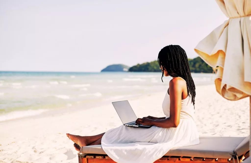 Black woman writing on laptop on her beach vacation. Keep reading to learn how to deal with traveling alone with anxiety on your solo trip