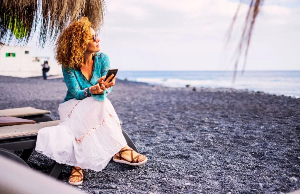 Mixed woman on the beach using the phone. Keep reading to learn how to deal with traveling alone with anxiety on your solo trip