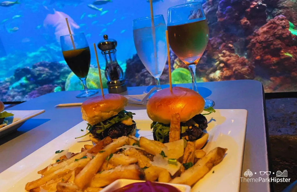 SeaWorld Orlando Resort Sharks Underwater Grill Kobe Beef Sliders with a glass of wine and water in front of panoramic views of the shark aquarium. Keep reading to learn more about Sharks Underwater Grill at SeaWorld Orlando.