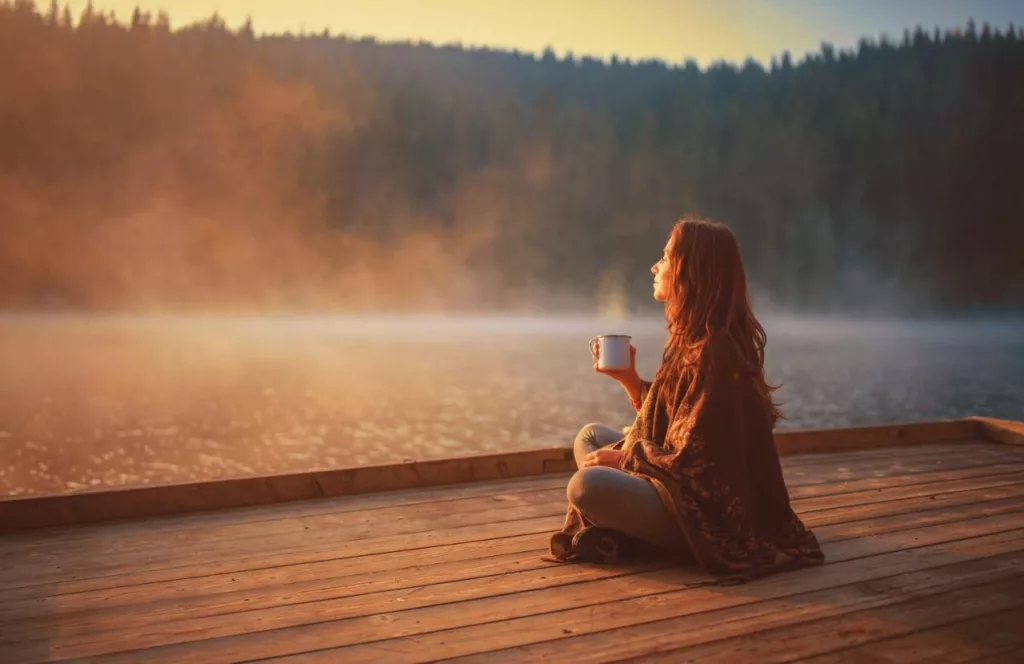 White woman on pier next to lake meditating with cup of coffee. Keep reading to learn how to deal with traveling alone with anxiety on your solo trip