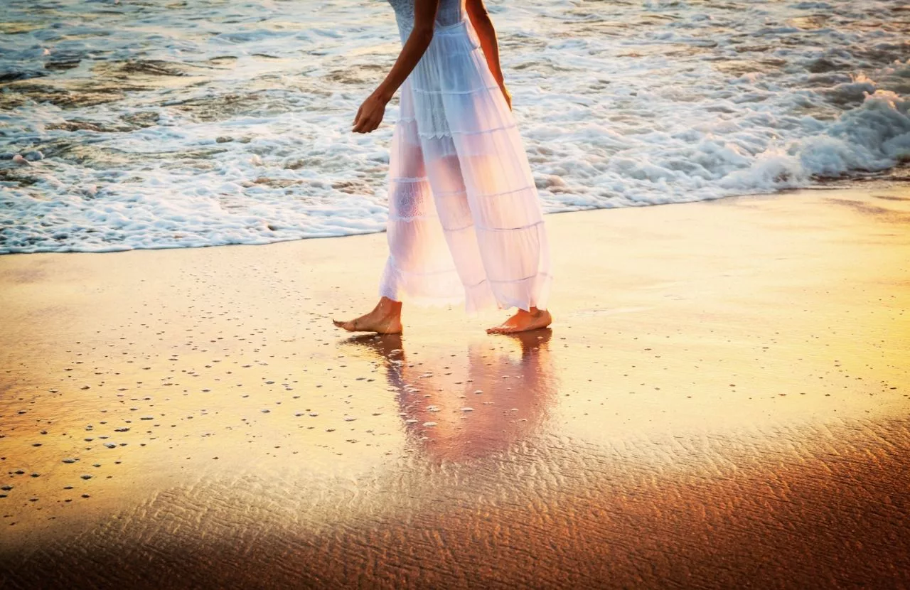 Woman with white dress walking on the beach sand next to water. Keep reading to learn how to deal with traveling alone with anxiety on your solo trip
